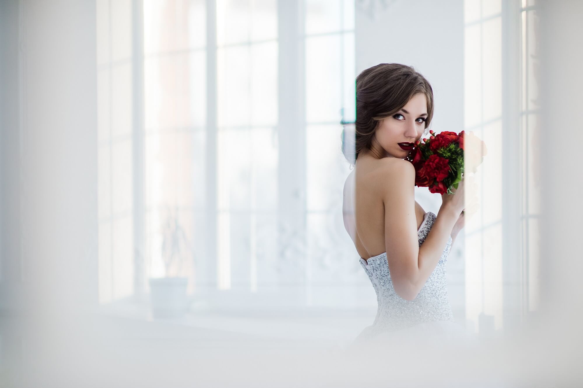 Glamorous young bride wearing wedding dress with flowers standing in front of doors indoors at luxury interior room.