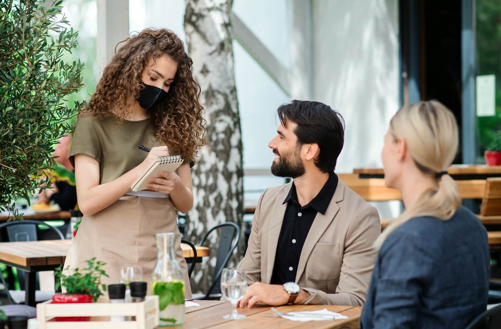 Woman waitress with face mask serving happy couple outdoors on terrace restaurant.