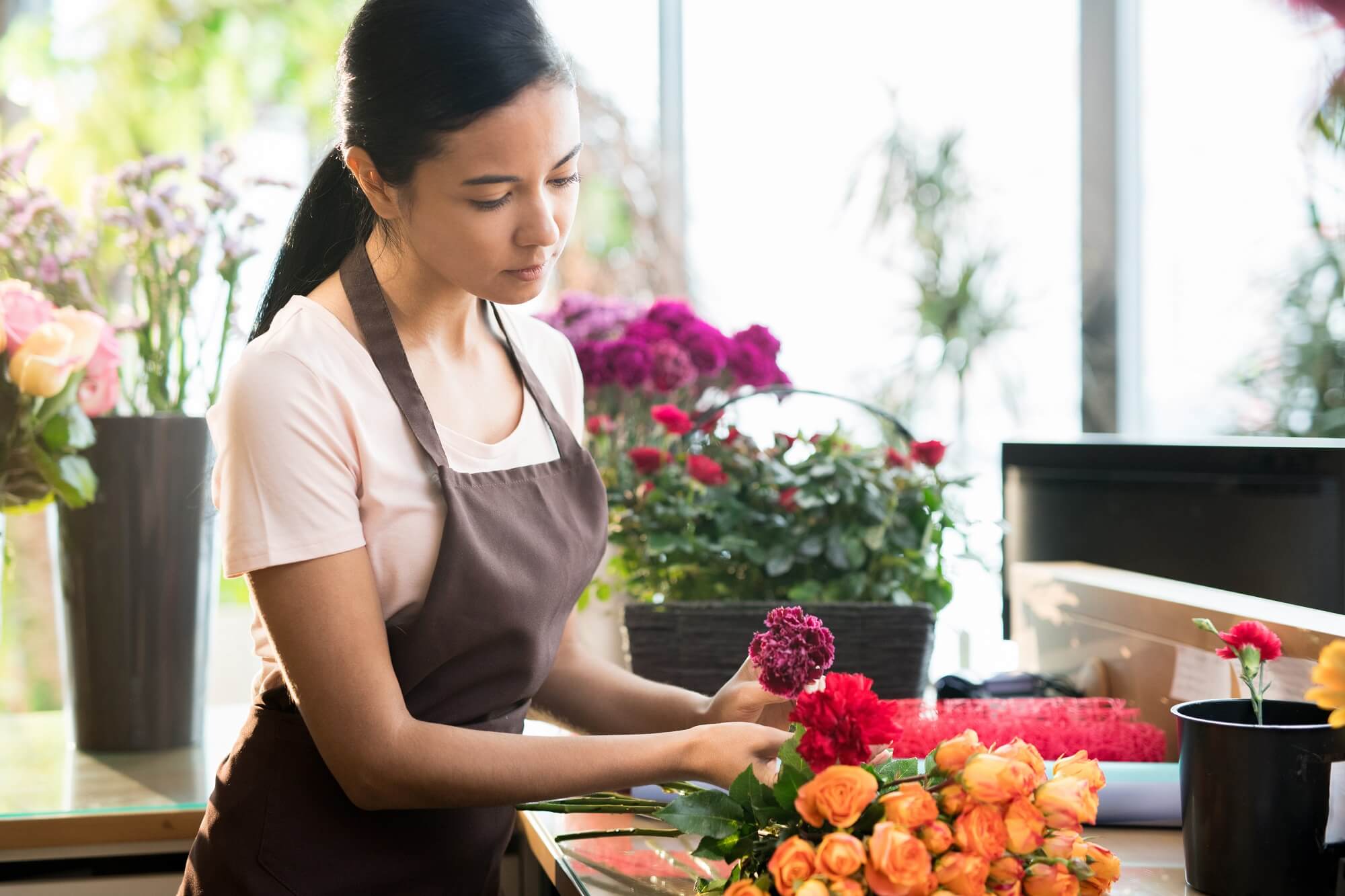 Young brunette florist in workwear sorting fresh roses and carnations on table during work in shop