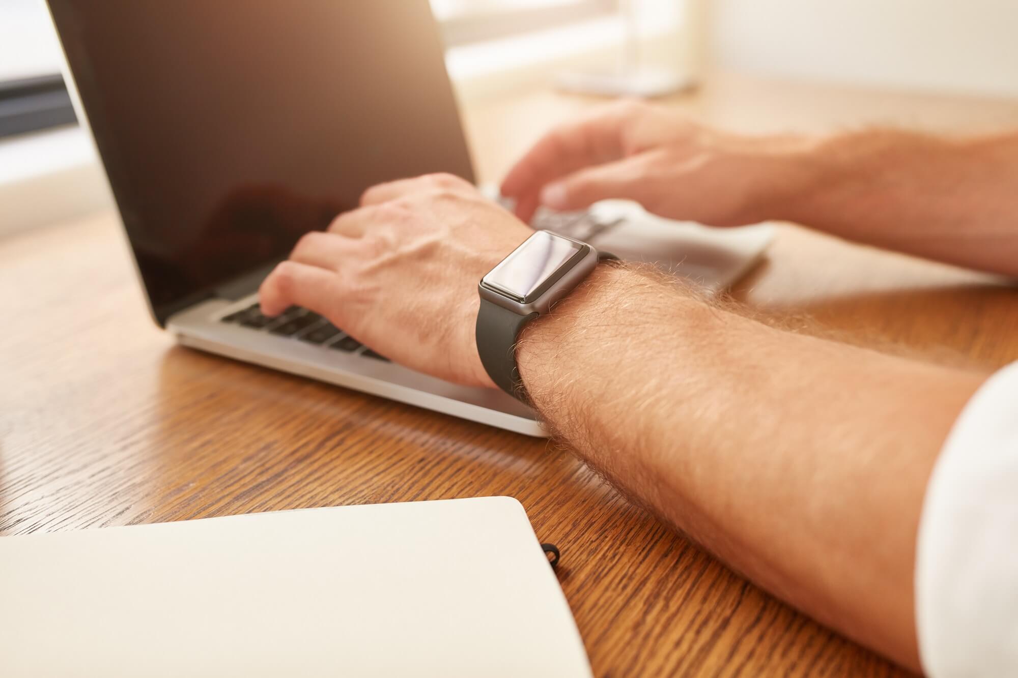 a man with a smartwatch working on a laptop while sitting at his desk.