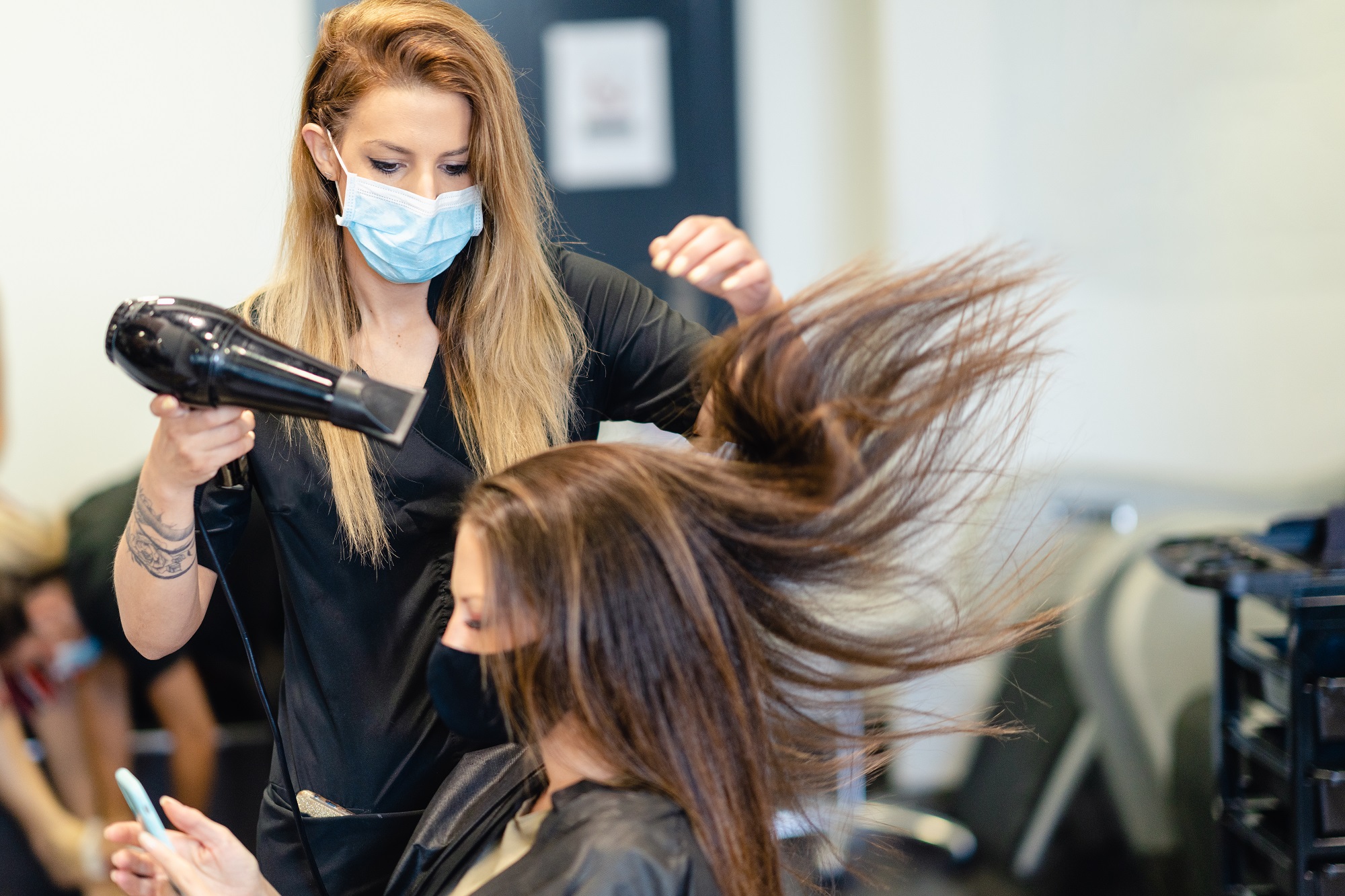 Female hairdresser drying her client's hair with a hairdryer, wearing protective masks 