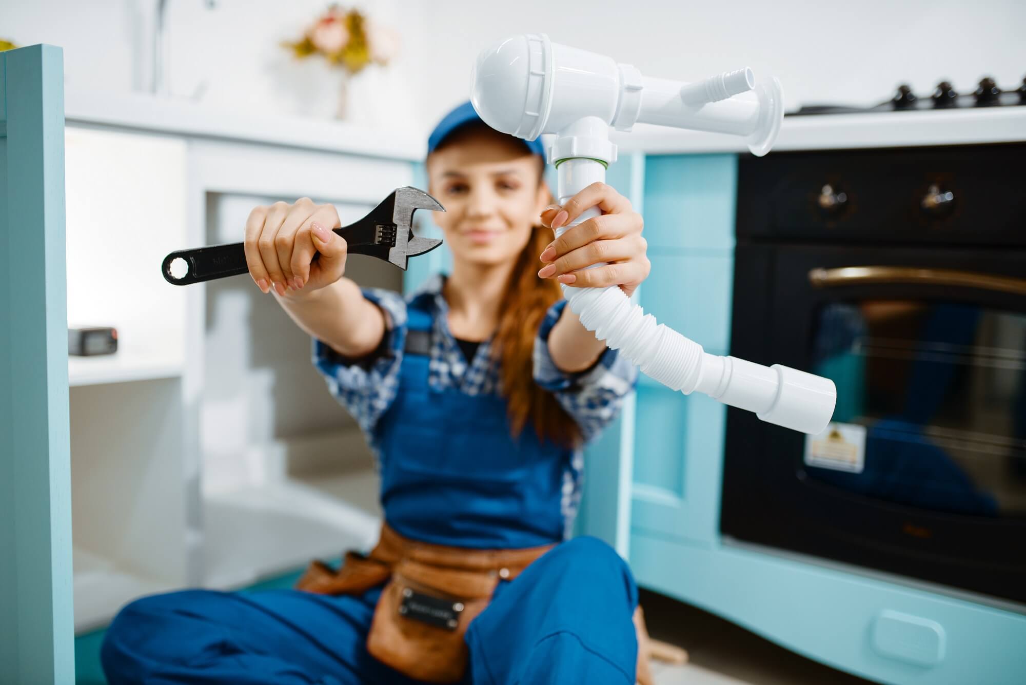 A young female plumber in uniform shows a wrench and pipe in the kitchen.